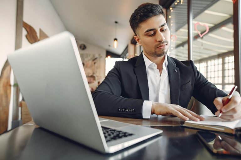 A young businessman of Indian origin wearing a suit takes notes while working on his laptop.