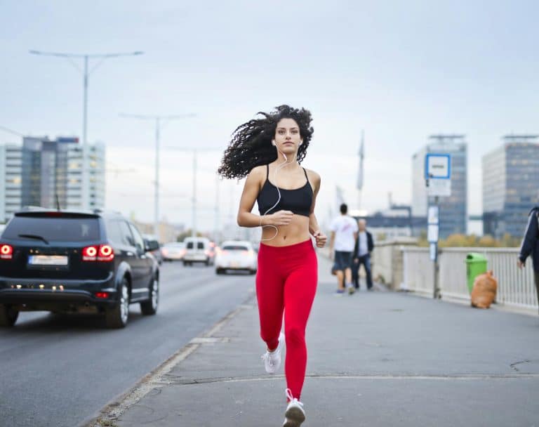 A young woman with curly hair wearing a black bra and red sports trousers is jogging down the street listening to music on her headphones.