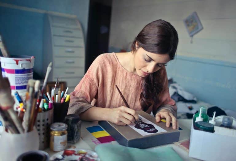 A young brunette woman in a pink blouse is painting on a frame in her studio. There are many pots of brushes around her.