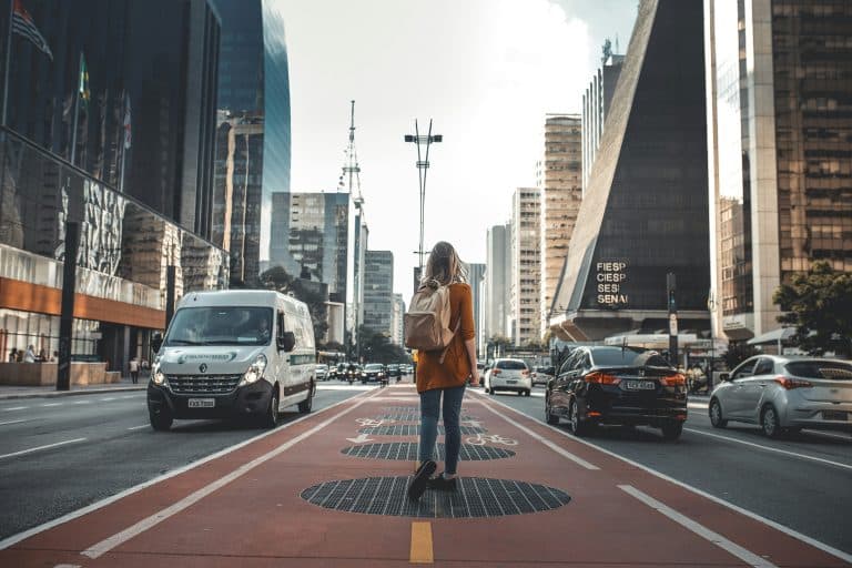 Photo of the back of a young woman carrying a rucksack. She is standing in the middle of a long street on a cycle path on Avenida Paulista in Brazil. Large buildings line the street and cars drive on both sides of the cycle path. The woman is looking towards the sky in the distance.