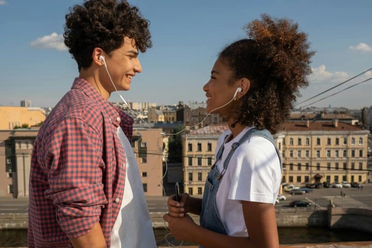 Two young people, a man and a woman, smile at each other and share the same headphones, listening to music on a phone held by the woman. The young man is wearing a red checked shirt and the young woman a white T-shirt with denim overalls. They are standing on an elevated terrace overlooking a city-centre avenue.
