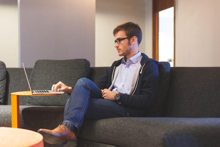 A young man wearing glasses is sitting on a grey sofa and consulting his laptop on a side table. The man is wearing a blue shirt, a hoodie and jeans.