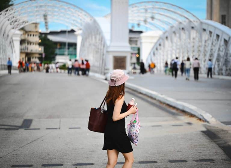 A young Asian woman seen from behind is wearing a short black dress, a pink bob hat and a brown leather handbag. She is standing on a bridge in Singapore, Asia.