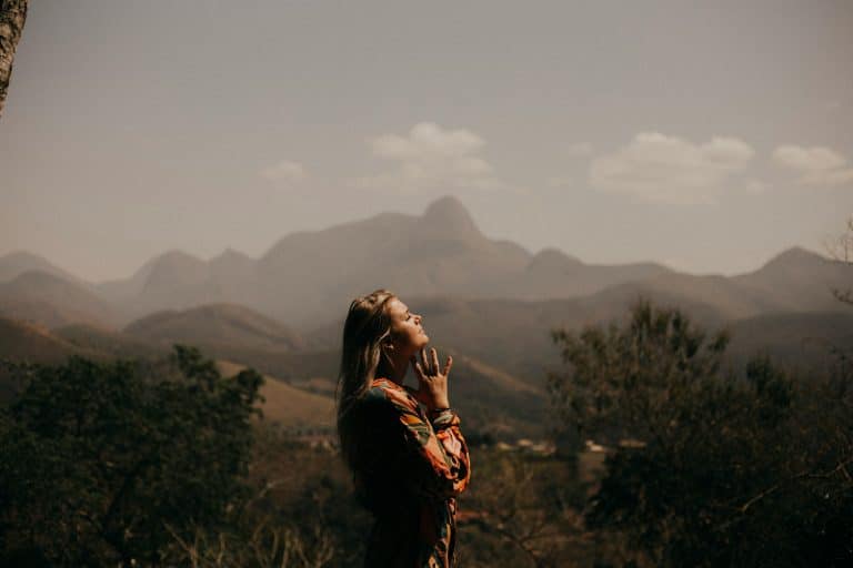 A woman closes her eyes and meditates outdoors, hands folded on her chin, in a natural setting in the mountains.