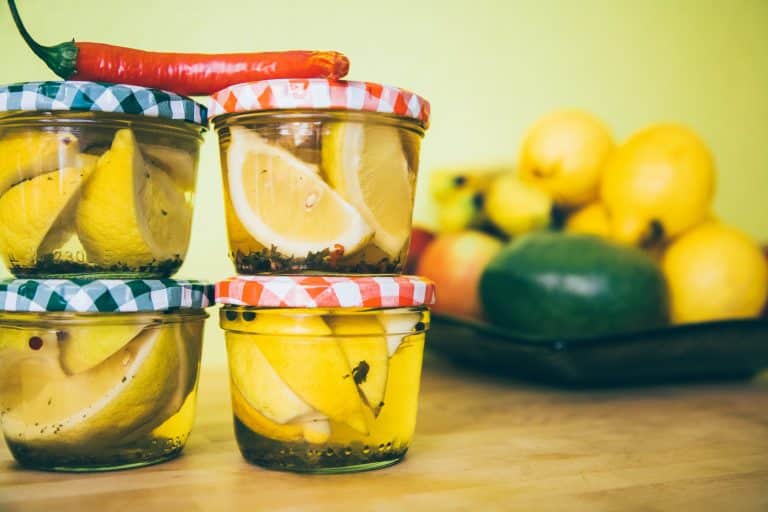 Lemons in marinade in glass jars stacked on top of each other. The background of the photo is also yellow and lemons and other fruits can be seen in a fruit basket in the background.