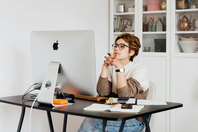 A woman with short hair and glasses crosses her fingers and looks intently at her computer screen. She is sitting at her desk and has been taking notes in her notebook and using a calculator.