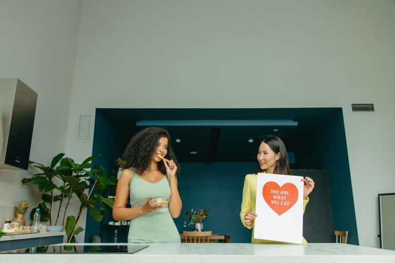 Two young women, one African and one Asian, are standing facing each other in front of a kitchen worktop. The kitchen walls are white and dark blue. One of the women is holding a sign that reads ‘You are what you eat’.